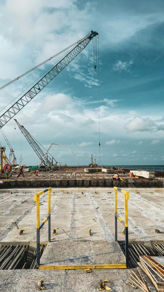 A bustling construction site by the sea with cranes and workers under a vibrant blue sky.